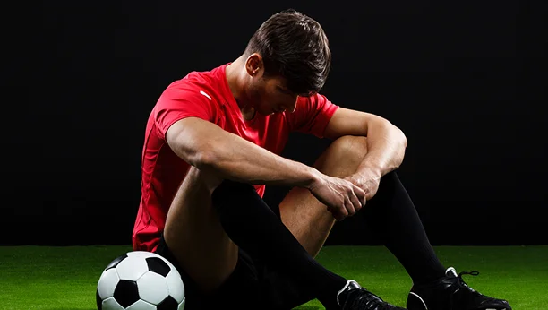 Un footballeur assis sur le terrain, la tête baissée, à côté d'un ballon, exprimant sa déception ou son sentiment de défaite après un match.